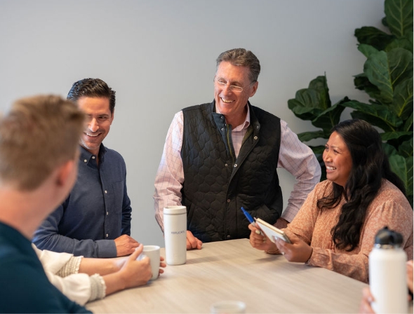 Four Replicate employees smile and converse around a table.