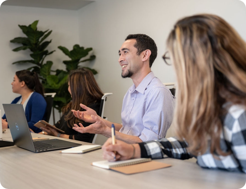 Four Replicate employees sit beside each other in a conference room. One employee is in focus, smiling and gesturing toward his open laptop.